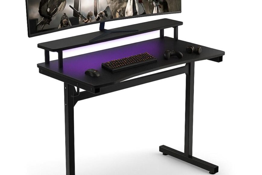Een modern gamingbureau met een breed scherm dat een actief spel toont, met een toetsenbord en een muis op de tafel. De onderkant van het bureau is verlicht met een paarse gloed, wat een stijlvolle uitstraling geeft.