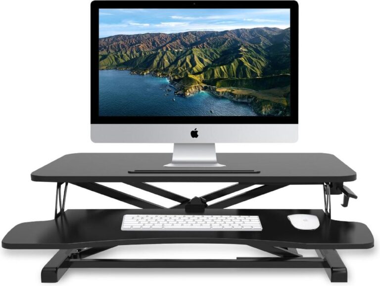 Een verstelbaar bureau met een iMac-computer bovenop en een toetsenbord en een muis op het lagere plankje. De achtergrond toont een prachtig uitzicht op bergen en water, wat een frisse en inspirerende werkomgeving suggereert.