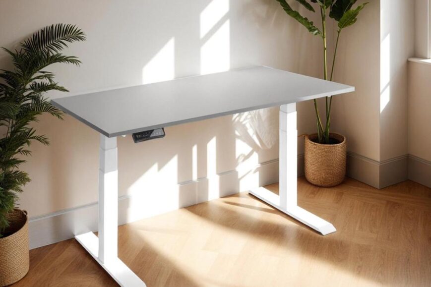 Een modern verstelbaar bureau van grijs en wit met een minimalistisch ontwerp, geplaatst in een lichte kamer met natuurlijk licht. Op de achtergrond zijn groene planten en ingelijste kunstwerken zichtbaar, wat de ruimte een frisse uitstraling geeft.