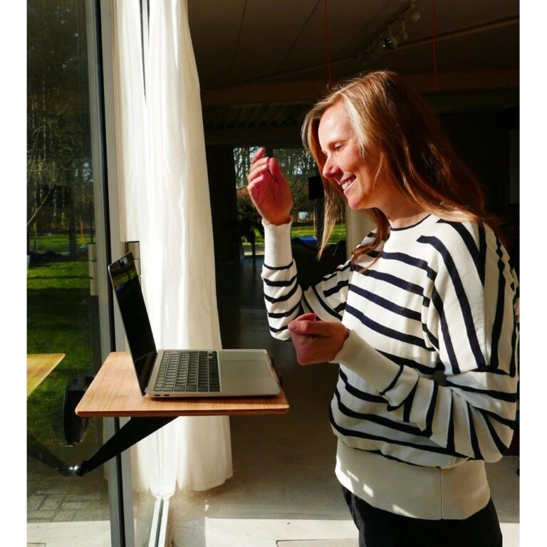 Vrouw met een gestreepte trui die enthousiast naar een laptop kijkt die op een venstertafel staat. Ze maakt een gebaar met haar hand terwijl ze glimlacht in een goed verlichte ruimte met uitzicht op een groene buitenomgeving.