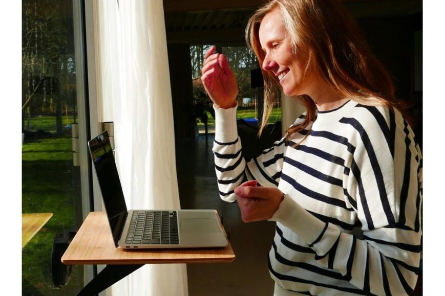 Vrouw met een gestreepte trui die enthousiast naar een laptop kijkt die op een venstertafel staat. Ze maakt een gebaar met haar hand terwijl ze glimlacht in een goed verlichte ruimte met uitzicht op een groene buitenomgeving.