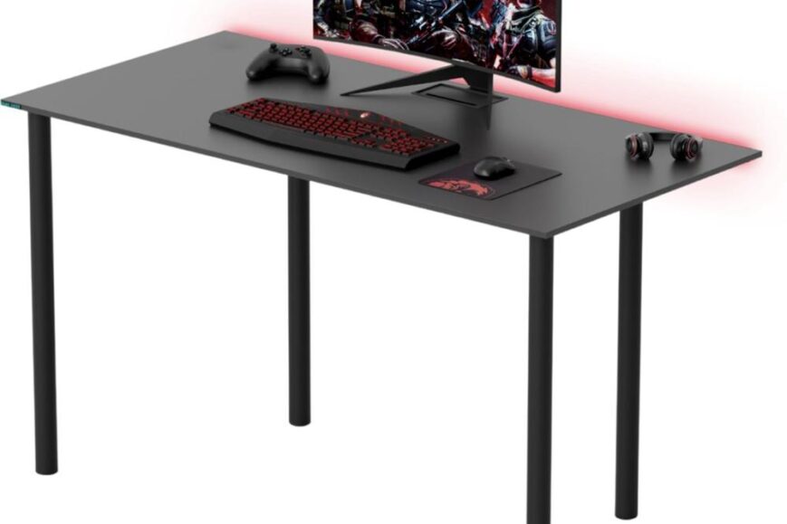 Een zwart bureau met een computer beeldscherm waarop een gaming afbeelding te zien is. Naast het scherm liggen een gaming toetsenbord, muis, een controller en een set hoofdtelefoons.