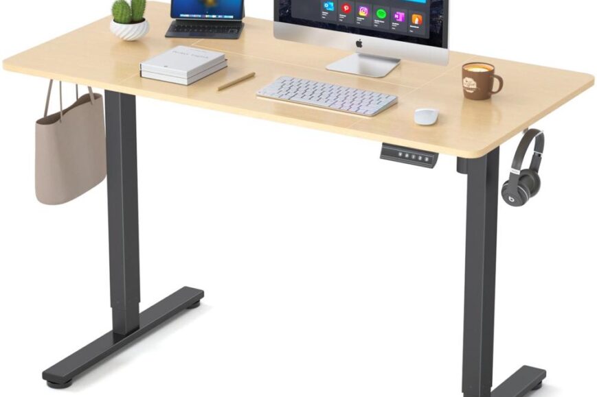 Een moderne bureauopstelling met een licht houten blad en zwarte onderstel, voorzien van een Apple desktopcomputer, een laptop, een toetsenbord, een muis en een mok koffie. Aan de zijkant hangt een tas en zijn er koptelefoon en een kleine cactus op het bureau geplaatst, wat een uitnodigende werkomgeving creëert.