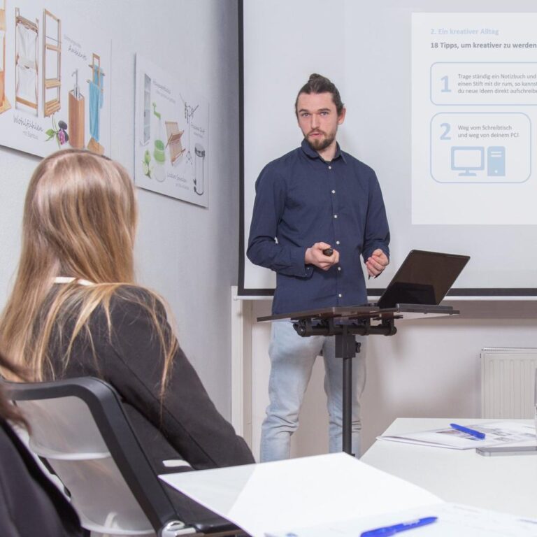 Een man presenteert tijdens een bijeenkomst met een laptop en een projector op de achtergrond. Het publiek bestaat uit enkele mensen die aandachtig naar de spreker luisteren.