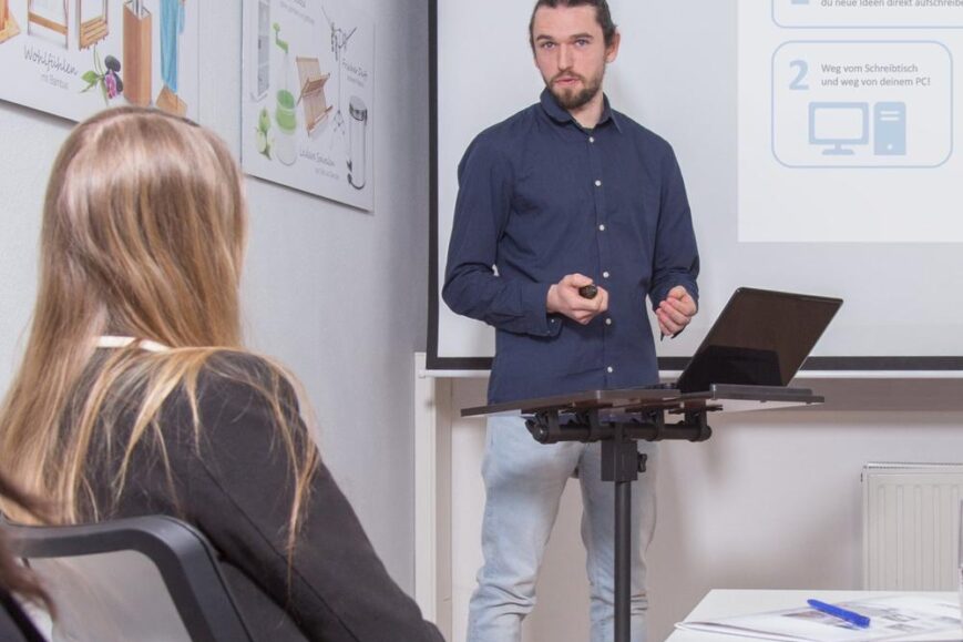 Een man presenteert tijdens een bijeenkomst met een laptop en een projector op de achtergrond. Het publiek bestaat uit enkele mensen die aandachtig naar de spreker luisteren.