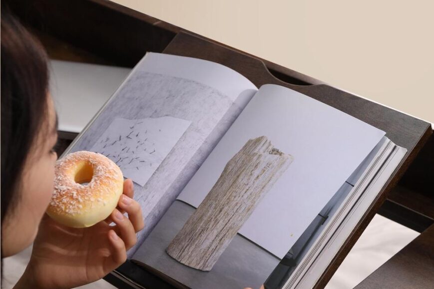 Een persoon leest een open boek terwijl hij een donut vasthoudt. De pagina's tonen een kunstwerk van een houten structuur en een abstracte afbeelding op een muur.